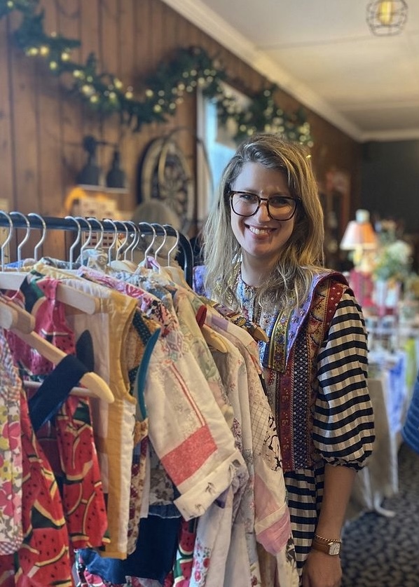 Woman with glasses smiling standing behind rail of handmade clothing